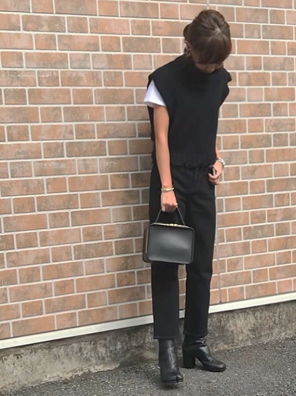 レンガ調の背景の前で黒のワントーンコーデで立つ女性
