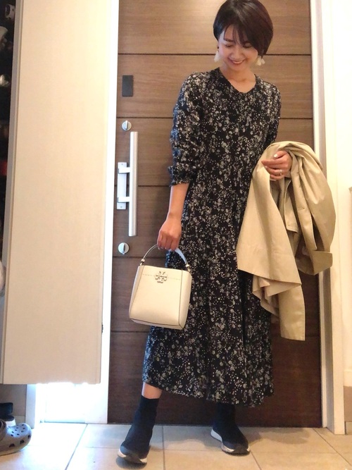 黒ベースの白の小花柄ワンピースに白のバッグとベージュのコートを手に持ち、黒のブーツを履いたコーデ