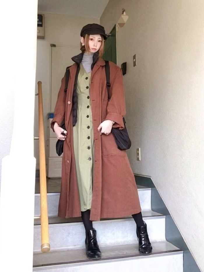グレーのハイネックトップスにカーキ色ワンピとブラウンのジャケットを羽織り、黒のダウンベストと黒のキャスケット、ショルダーバッグと黒のブーティをコーディネート